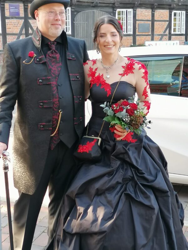 Brautpaar im Steampunk Stil mit schwarzem Brautkleid und roter Spitze vor Fachwerkhaus – VeryNice Brautmode Winsen