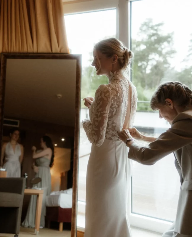 Braut beim Ankleiden mit Spitzenkleid und Stehkragen am Fenster – VeryNice Brautmode Winsen