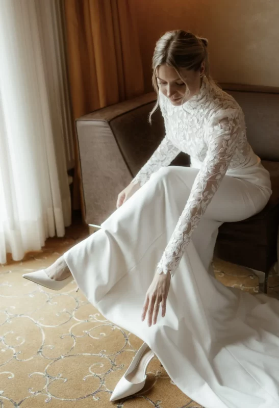 Braut in Spitzenkleid mit Stehkragen sitzend im Hotelzimmer – VeryNice Brautmode Winsen