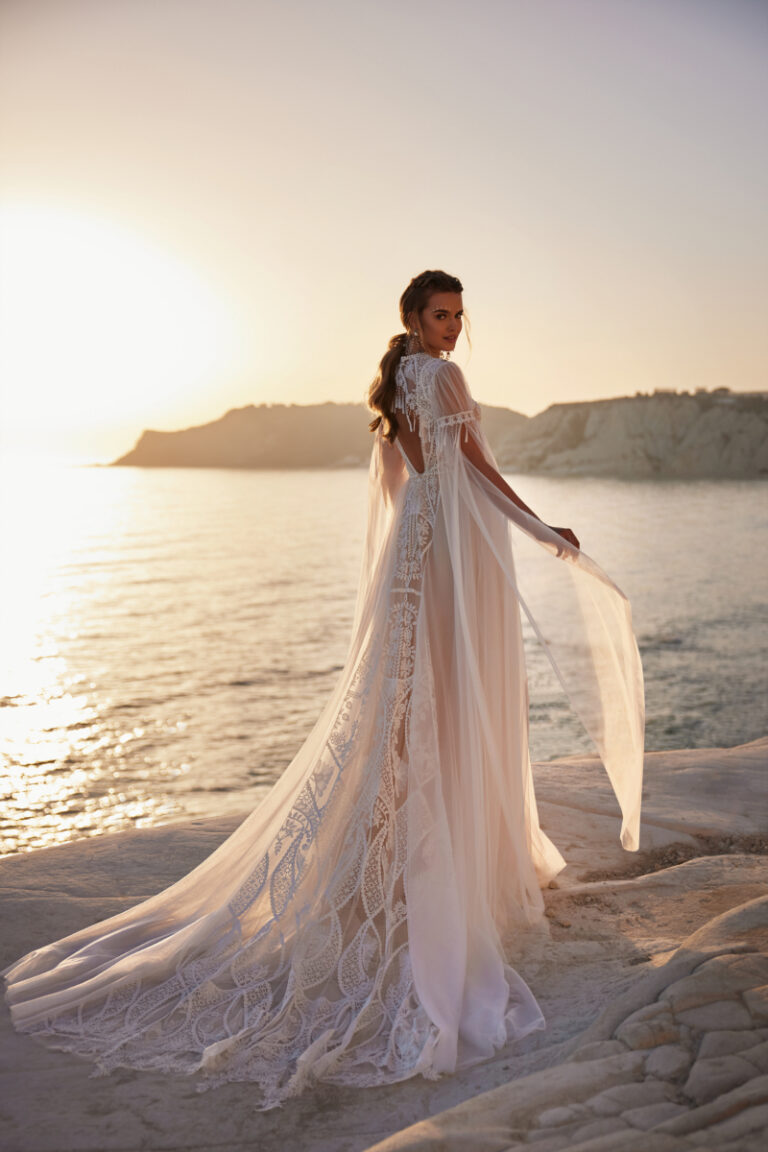 ANWAR Brautkleid von Dama Couture mit wehendem Schulter-Cape und Boho-Spitze – Brautmode, Rückenansicht an felsiger Küste