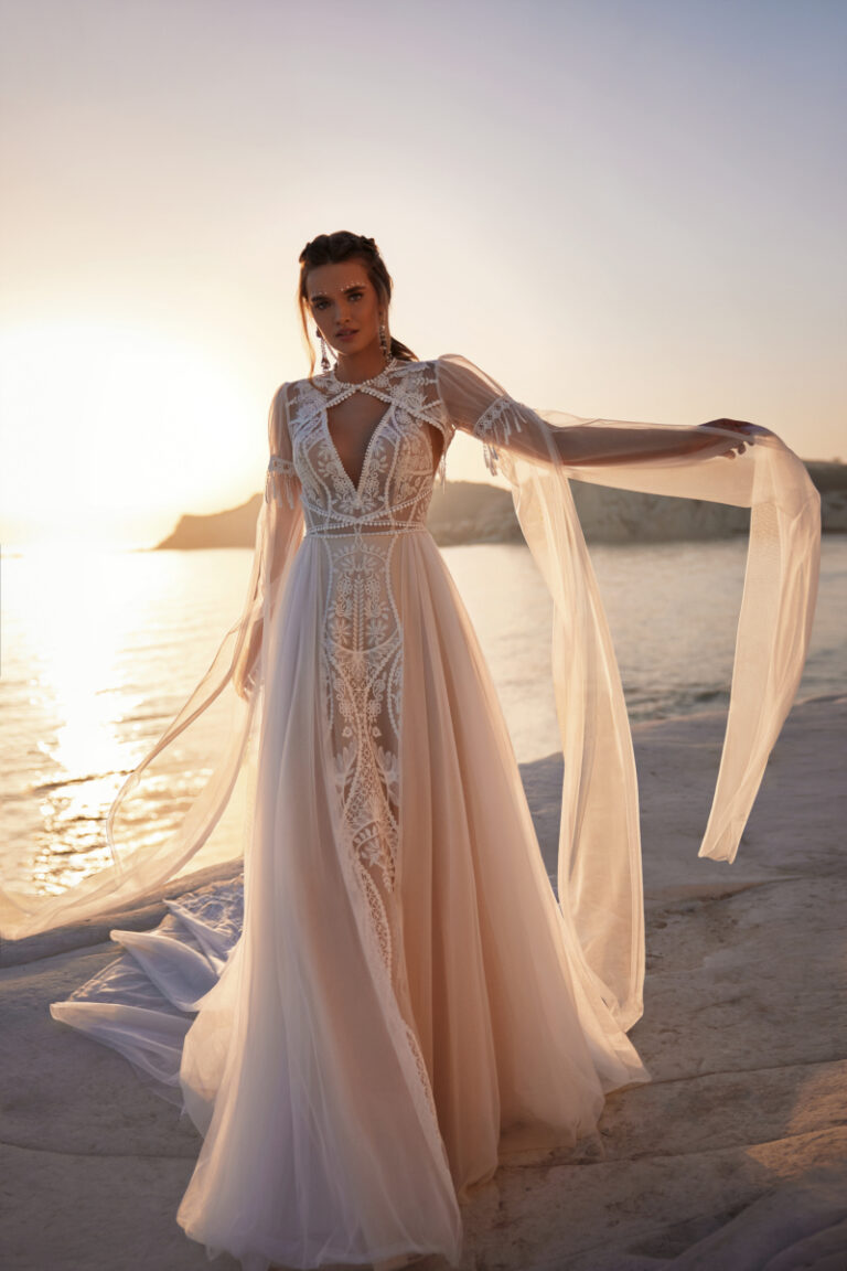 ANWAR Brautkleid von Dama Couture im Boho-Stil mit Schulter-Cape – Brautmode, Braut mit ausgebreiteten Armen am Strand