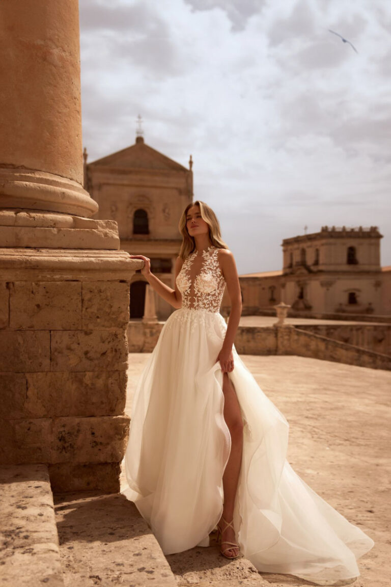 BANTA Brautkleid mit Illusionsspitze und Beinschlitz – Brautmode Winsen, Braut an Steinsaeule vor Altstadt-Kulisse