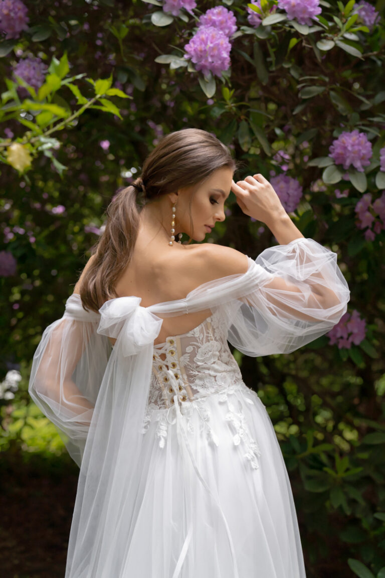 Boho-Brautkleid Lanka von Dama Couture mit Wasserfall-Ausschnitt und weiten Ärmeln – Brautmode, Braut vor blühendem Rhododendron