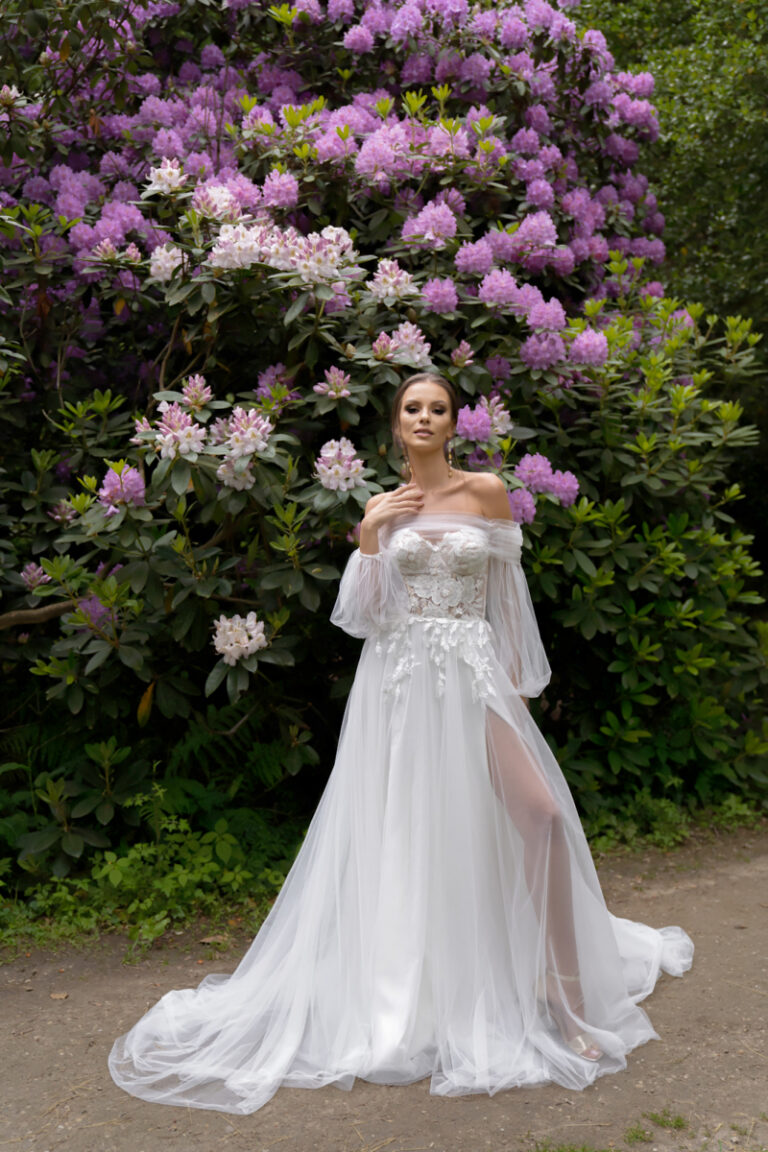 Boho-Brautkleid Lanka von Dama Couture mit Beinschlitz und langer Tuellschleppe – Brautmode, Ganzkörperansicht im Blumengarten