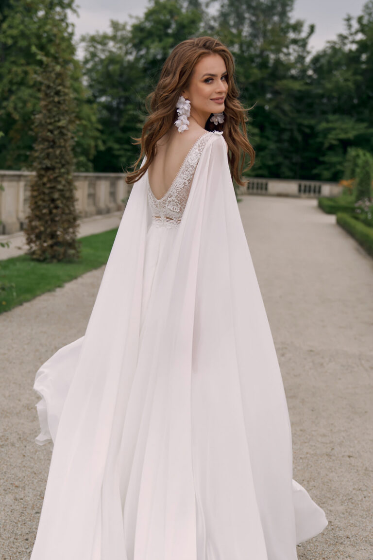 Brautkleid Atena von Dama Couture mit Schulter-Cape aus Crepe-Chiffon – Brautmode, Rückenansicht im Schlosspark mit wehendem Umhang
