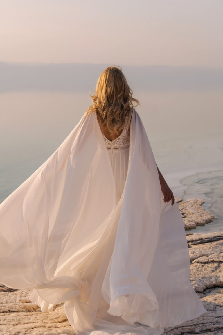 Brautkleid Atena von Dama Couture mit wehendem Schulter-Cape – Brautmode, Rückenansicht am felsigen Ufer