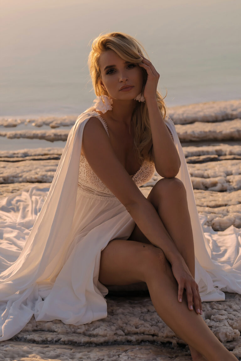Brautkleid Atena von Dama Couture mit Schulter-Cape aus Crepe-Chiffon und Spitzenoberteil – Brautmode, Braut sitzend am Strand bei Sonnenuntergang