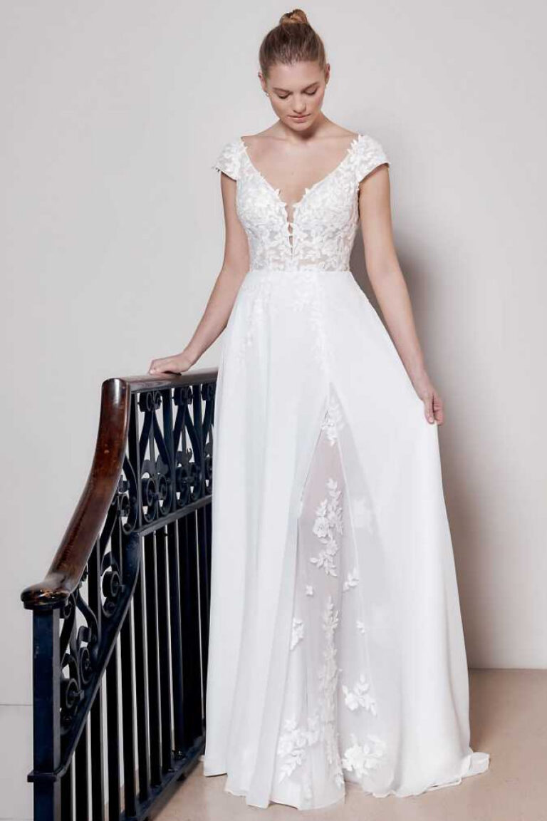 FRIDA Brautkleid mit tiefem V-Ausschnitt und Beinschlitz mit Spitzenapplikation – Brautmode Winsen, Vorderansicht an Treppengeländer