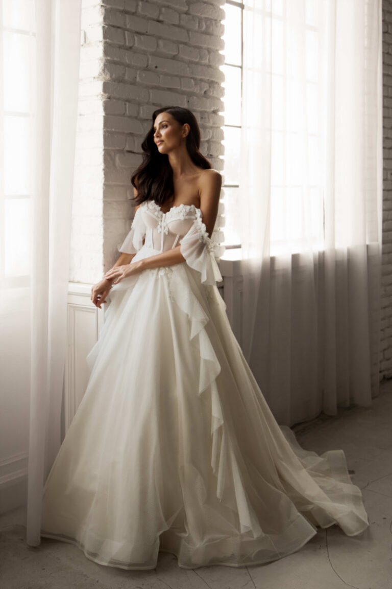 ZAYANA Brautkleid mit Carmen-Ausschnitt und Rüschenärmeln – Brautmode Hamburg, Braut am Fenster in bauschigem Tuellrock